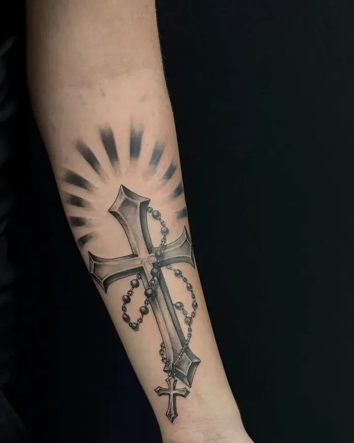 Black and grey cross with a rosary and light rays, on a forearm